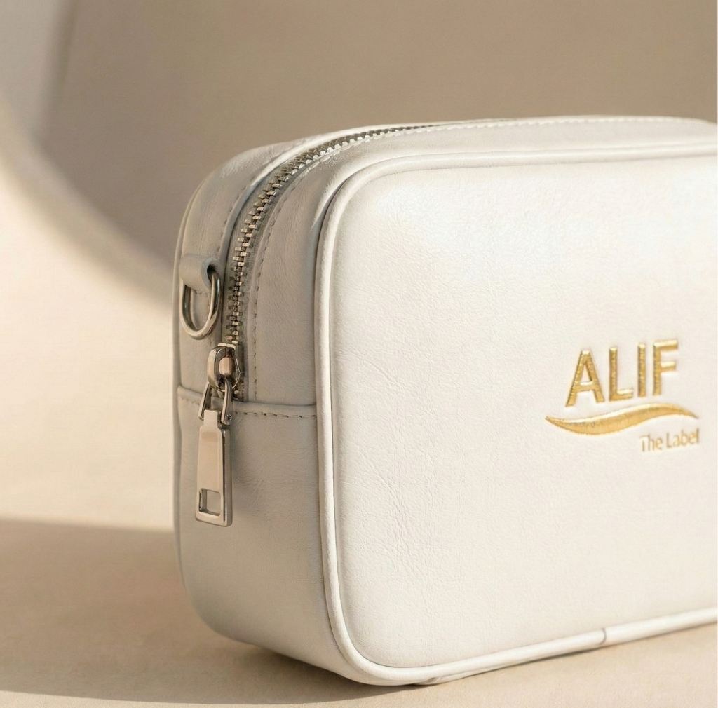 White downttown sling bag with gold zipper and 'ALIF The Label' logo on a beige background